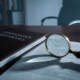 Gebarsten vergrootglas op open compliance-documentmap onthult kleine lettertjes, koele kantoorverlichting werpt schaduwen