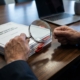 compliance-documenten-controle-audit-zakelijkjpg - Hoek en Blok.IT Handen met vergrootglas onthullen hiaat tussen compliance-documenten op houten vergadertafel met rode tabbladen