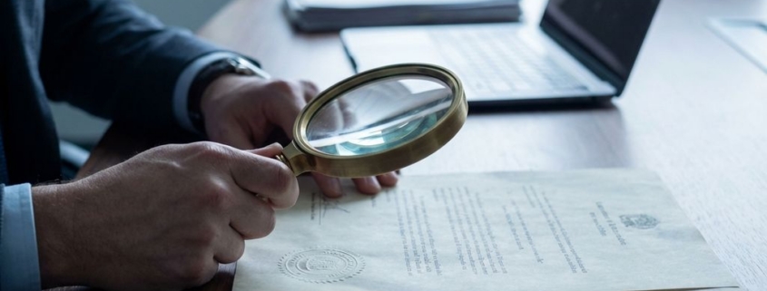 document-verificatie-vergrootglas-zakelijk-kantoorjpg - Hoek en Blok.IT Zakelijke handen houden vergrootglas boven officieel document met zegel op donkere houten vergadertafel
